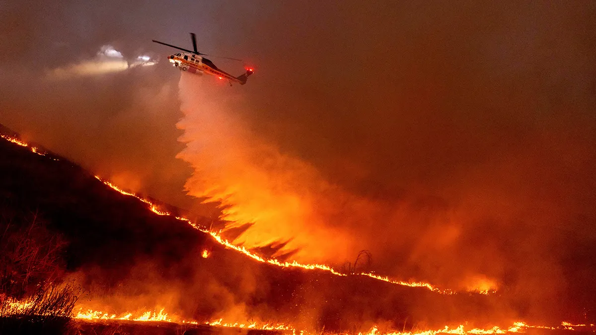 Wildfire burning across hillside terrain above a populated area, with smoke rising into the sky